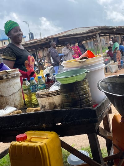 A woman wearing a green headwrap and a black shirt with gold embellishments smiles broadly at the camera in what appears to be an outdoor market. She is positioned in the foreground, with a wooden table laden with various containers and bottles in front of her. The table holds large plastic buckets, several bottles of yellow liquid, a dark red bottle, and metal containers. In the background, market stalls with corrugated metal roofs are visible, with other people milling about. The scene suggests a bustling marketplace environment, likely in a rural or semi-urban setting in Wang, Nigeria, given the presence of the woman and the style of the market stalls. The lighting suggests it is daytime, with an overcast sky.
