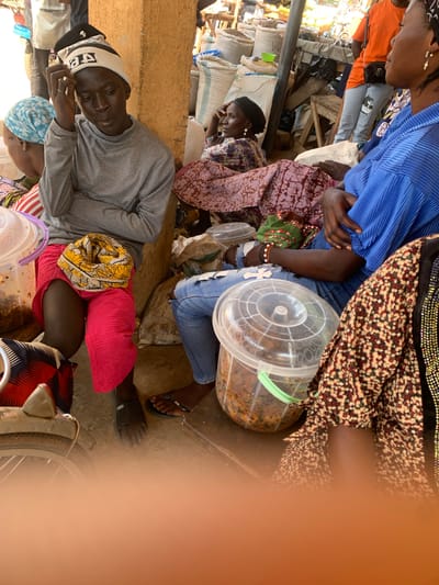In Kafanchan B, Nigeria, a street market scene is captured. In the foreground, a young person wearing a gray long-sleeved shirt and a beanie hat with black and white stripes sits with their legs crossed, one foot bare and the other in a black sandal. A yellow and white patterned scarf is tied around their waist. To the left, a plastic container filled with food is visible, next to a bicycle wheel and frame. Further to the left, a person in a blue headscarf is partially visible, with their back to the camera.
In the mid-ground, two women are seated. One, on the left, wears a dark headscarf and a patterned dress, her right hand resting on her chin. The other woman, in the center, wears a pink and white patterned dress and a dark headscarf.
In the background, a person in an orange shirt and light blue jeans with a cat patch is standing. Bags of what appear to be grains or other market goods are stacked behind them, and wooden stalls are visible. The ground is dusty and uneven, consistent with an outdoor market environment. The overall lighting suggests it is daytime.