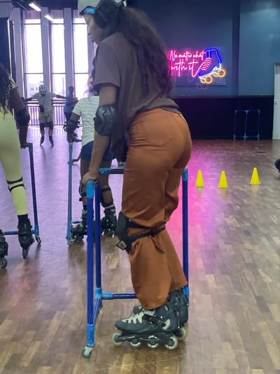 The image shows a group of people, including a woman wearing a brown jumpsuit and inline skates, practicing inline skating at what appears to be an indoor skating rink. The woman is holding onto a blue support structure, presumably for balance. Other individuals can be seen further in the background, some also with roller skates and protective gear. There are some yellow cones in the background, and a neon sign on the wall. The flooring is wooden. The location is difficult to ascertain as Moba is a large city in Nigeria, but the presence of an indoor skating rink suggests it is an urban area.