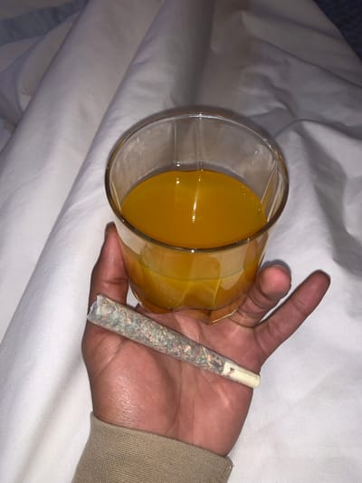A hand holds a glass of orange juice and a rolled cigarette. The juice is in a clear glass with faceted sides. The cigarette has a white filter. The hand is dark-skinned and partially covered by a beige-colored sleeve. The background is a rumpled white sheet or blanket.