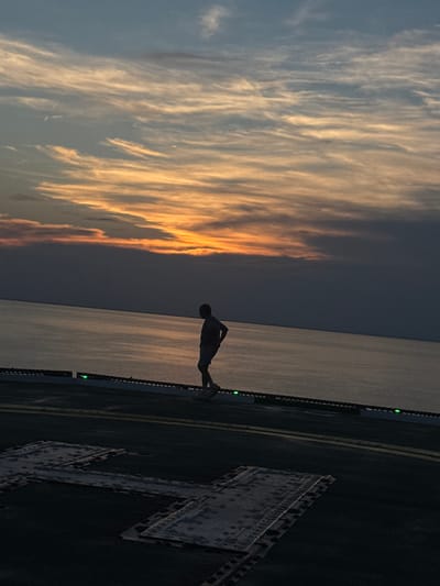A silhouetted figure stands on a helideck at sunset. The sky is a canvas of streaky clouds, with hues of orange and yellow bleeding into a dusky blue. The sea stretches out behind the figure, reflecting the warm tones of the sky. The helideck itself is dark, with yellow lines marking its surface and a large, metallic "H" symbol painted on it. Green lights are visible along the edge of the helideck. The scene evokes a sense of peace and solitude, as the individual appears to be contemplating the vastness of the ocean and the beauty of the sunset. The time of day is clearly sunset due to the dramatic colors in the sky and the low angle of the light. The location is unknown, but the presence of a helideck suggests it might be a ship, an oil rig, or a remote landing area.