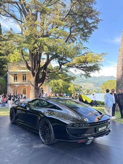 The image shows a black Brabus car on display at an event in Como, Italy. The car is parked on a dark surface, likely a platform for display. In the background, there is a large tree, a building with a yellow facade, a crowd of people, and a view of the lake and mountains. Some people are observing the car and the surroundings. The sky is blue with some clouds. The angle is slightly rotated, and the car is in focus.