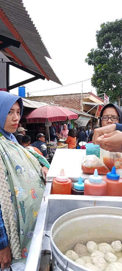 The scene takes place in a market in Kecamatan Kutawaringin, Indonesia, at latitude -6.973 and longitude 107.52. A woman wearing a blue headscarf and a green and white patterned shawl stands at a food stall, looking at the camera. In front of her is a metal counter with a pot of meatball soup. The pot is surrounded by containers filled with chili sauces and other condiments. Several other people are visible in the background, some are buying food, others are walking by. There are buildings and trees in the background. The stall is covered by a corrugated metal roof. The scene appears to be on a busy street, with a lot of pedestrian activity. The woman on the left is holding a bag in her left hand.  She is looking at the camera. There is a man standing behind her, holding his phone. He is wearing a white shirt and a blue cap.