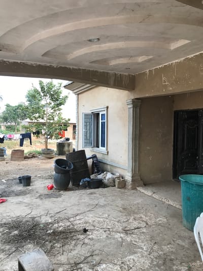 The image depicts a building exterior with a cream-colored facade. A decorative arched ceiling overhangs, featuring recessed circular elements. A window with a blue frame is visible below. Various items are scattered on the ground, including plastic bins, pots, wood, and what appear to be discarded plant debris. Laundry is hanging on a clothesline in the background with trees around it.