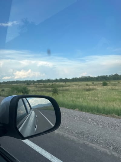 The image captures a view from the passenger side of a vehicle driving on a paved road, which appears to be in or around Sayanogorsk, Russia. In the foreground, a portion of the car's side mirror is visible, reflecting the road behind and the sky. The road is gray and appears to have a white line marking the edge. To the right of the road, a grassy field stretches into the distance, dotted with scattered bushes and a line of trees in the background. The sky is a vibrant blue with white, wispy clouds. The overall scene depicts a rural or semi-rural landscape, possibly on the outskirts of Sayanogorsk, given the context of the location. There are no people visible in the image.
