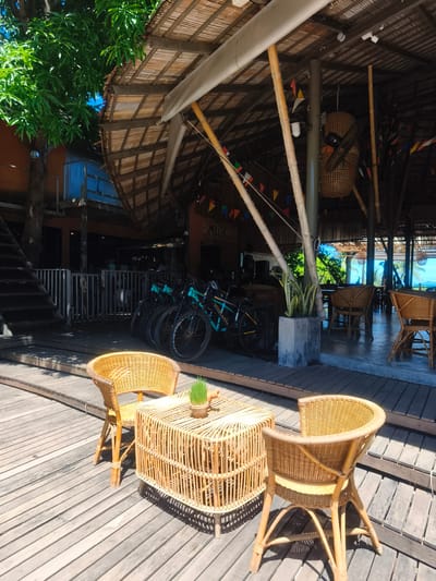 This media file captures an outdoor seating area at Baan Thong Po, Thailand, likely during the daytime under clear skies. The scene is set on a wooden deck. In the foreground, two woven wicker chairs and a matching coffee table are arranged. A small potted plant with green grass sits on the table, adding a touch of nature.
The background reveals a covered structure with a thatched bamboo roof supported by bamboo poles. Colorful triangular flags are strung along the ceiling, suggesting a festive or casual atmosphere. Beyond this open-air area, bicycles are parked, hinting at activities like cycling tours or rentals. Further in the distance, through openings in the structure, palm trees and the bright blue sky are visible, indicating a tropical environment. The overall impression is one of a relaxed, tropical resort or cafe setting. No people are visible in the frame, and no text is discernable.