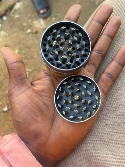 This image depicts a dark-skinned human hand, positioned palm-up, holding two disassembled components of a metallic grinder. The hand rests on a light brown, woven fabric garment, with a pink textile sleeve visible near the wrist. Both circular grinder components feature an array of diamond-shaped teeth and circular perforations. Significant residue of greenish-brown plant material, consistent with cannabis, is visible adhering to the teeth and internal surfaces of both components. The upper grinder component exhibits a decorative gold-colored band on its exterior. The background consists of an unfocused outdoor area with dirt or sandy ground. A blurred footwear item, consistent with a sandal, is partially visible in the upper left background. The scene is located in Katon Rikkos, Nigeria.