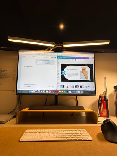 This media file captures a late-night desk setup in Santa Clara Ocoyucan, Mexico, suggesting focused work or study. The central subject is a large computer monitor, positioned vertically (portrait mode), displaying two active applications.
The top application is Canva, showing a presentation slide titled "Epidemiología" (Epidemiology). The slide features an image of a person's neck and chest, along with Spanish text discussing epidemiology and two light blue snowflake/starburst graphics. The top bar of this application indicates "Mon 27 Apr 11:36 p.m." and "U UPAEP," likely referencing a university.
Below, the monitor displays Google Gemini, an AI assistant. This window contains Spanish text about "Fisiopatología de Laringomalacia: Esquema Visual" (Pathophysiology of Laryngomalacia: Visual Scheme), detailing the structural weakness in the larynx. A prompt reads, "Introduce una petición para Gemini" (Introduce a request for Gemini). The monitor's dock, visible at the bottom, shows various application icons including Safari, Mail, Calendar (with '27'), and Notes.
Two long, warm-toned LED light bars are attached to the top of the monitor, illuminating the workspace. To the right of the monitor, on a light brown, textured desk mat, sits a white Apple-style keyboard and a black ergonomic Logitech mouse. The monitor rests on a simple, light wood-colored riser. In the upper right background, a pencil holder filled with pens and pencils, along with a ruler, is visible. The overall scene suggests a dedicated study or work session, likely for academic or professional purposes given the content on screen, occurring late in the evening.