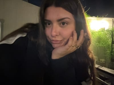A woman with long brown hair and fair skin is facing the camera. Her hand is near her face, displaying several rings on her fingers. She wears a dark top with a glimpse of a white collar. The background shows a dimly lit outdoor space, including greenery illuminated by a light, suggesting a nighttime setting. The photo is slightly tilted.