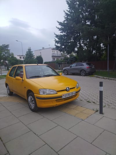 The image depicts a street scene in Poznan, Poland. A bright yellow Peugeot 106 is parked on a paved sidewalk, facing left. The car's license plate is visible and reads "BY 0170 EB-4". Behind the Peugeot, a grey Audi Q3 is parked on the street. The environment includes trees, buildings, and a lamppost. The sky is overcast.