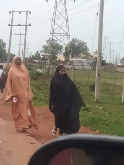The image shows two women walking outdoors in Kassa Market, Nigeria. The woman on the left is wearing a light orange hijab and dress, and the woman on the right is wearing a black hijab and dress. Both women are walking on a dirt path. The background shows a grassy area with trees, buildings, and several utility poles with power lines. The sky is overcast, suggesting it might be a cloudy day. The scene appears to be during daylight hours. There is some visible text on a sign in the background, which appears to read "ZIRA". The overall mood of the image is neutral, depicting a common scene in an outdoor market.