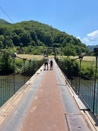 A man and a child are walking across a suspension bridge over a river. The bridge is made of metal and wood with a metal railing. The man is wearing a t-shirt and shorts, and the child is wearing a hat and shorts. They are walking away from the camera towards a wooded hill. The bridge is located in Moračko Trebaljevo, Montenegro. The environment is lush and green with trees and hills. The sky is clear and blue.