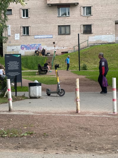 This media file, captured in Kuzmolovskiy, Russia, depicts an outdoor scene with several individuals and objects. The setting appears to be a residential area with a brick building featuring multiple windows and a grassy area adjacent to a paved walkway.
In the foreground, several people are present. One person in a blue shirt and white cap stands near a scooter. Further down the walkway, a person in dark clothing is leaning over. Another individual in a purple and dark jacket is walking away from the camera. In the background, a group of people are gathered, some sitting on a bench and some standing, with a couple of dogs.
Objects visible include a yellow and black scooter, a concrete trash receptacle, and a signpost with Cyrillic text. The text on the signpost indicates "Благоустройство общественной территории вдоль домов 24, 26 по ул. Железнодорожная в ГП Кузьмоловский Всеволожского района Ленинградской области," which translates to "Improvement of public territory along houses 24, 26 on Zheleznodorozhnaya Street in the urban settlement Kuzmolovskiy, Vsevolozhsky district, Leningrad region." Another sign above the entrance to a building reads "ПАРИКМАХЕРСКАЯ," meaning "Hairdresser's."
The sky suggests it is daytime, and the lighting indicates overcast conditions. The overall atmosphere seems casual, with people engaged in everyday activities.