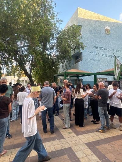 The image depicts an outdoor gathering of people in front of a light blue building with a textured facade. The building has a yellow textured roofline and features a crest with text in Hebrew. Below the crest, there is a large inscription in Hebrew. A green and white flag is attached to the building. The ground is paved with square tiles in shades of brown and beige. Several trees with green foliage are visible.

A group of people, appearing to be of various ages, are standing and interacting. Some individuals are holding plates or food. One person in the foreground is wearing a yellow paper crown. The gathering seems informal, with people dressed in casual clothing such as t-shirts, jeans, and shorts. The overall atmosphere appears to be a community event or celebration. The building's inscription and crest suggest the location is Givatayim, Israel.
