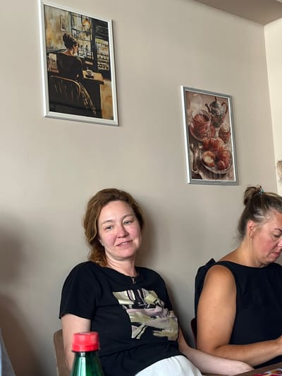 The photograph shows two women seated at tables in what appears to be a cafe in Donji Pobori, Montenegro. The woman on the left, with shoulder-length reddish-brown hair, is wearing a black t-shirt with a graphic design and white pants. She is looking slightly to her right, with a faint smile. The woman on the right, with her hair pulled back in a bun, is wearing a black sleeveless top. She is looking down at something on the table in front of her. Two framed pictures hang on the wall behind them. The picture on the left depicts a woman sitting at a table in a cafe, seen from behind. The picture on the right shows a tea set, croissants, and a bowl of sugar. A green bottle with a red cap is partially visible in the foreground on the left. The lighting suggests it is daytime, and the environment is indoors.