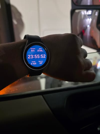 A person's wrist is shown wearing a smartwatch with a blue display. The watch face shows the time as 23:55:52, with additional information about battery (64%), date (27.06), steps (9425), and temperature (29°). The background is blurred, but appears to be inside a vehicle, possibly at night, with reflections of lights visible in what appears to be a side mirror. The person's hand is clenched. There is no information in the image to suggest a specific location in Frankfurt, Germany.