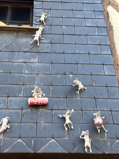 In Honfleur, France, a building's facade is adorned with numerous small, white, mouse-like figures. These figures are in various poses, some appearing to climb the slate-tiled wall, while others engage in activities. One group of figures is positioned on top of a red Coca-Cola can. Another figure is playing a trumpet, and one is depicted as a drummer, using a can similar to the Coca-Cola can as a drum. A figure is shown holding a small, colorful object, possibly a flag or a toy. The building has a dark-paned window on the upper left and a rustic wooden beam on the right. The slate tiles are dark gray and show some signs of weathering.