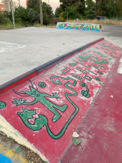 This image depicts a skateboard park in Badalona, Spain, with a focus on a red ramp adorned with green drawings. The drawings appear to be abstract figures, possibly resembling a stylized animal or creature with a long tail, and various geometric shapes including circles, squares, and lines. Some of the shapes are arranged in a manner that suggests letters or symbols. The ramp has a black coping along its edge. In the background, a larger skate feature with graffiti is visible, along with trees and buildings. The ground is concrete, with some scattered leaves and dust.
