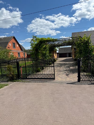 In Dovhe, Ukraine, a paved driveway leads to a property featuring a house and a garage. The house, constructed from red brick, has a dark-tiled roof and white decorative elements. Adjacent to it, a yellow building with a similar dark roof is visible. The driveway is partially covered by a metal pergola with climbing plants, casting dappled shadows on the ground. A black metal gate, partially open, guards the entrance to the property. The scene is set against a bright blue sky dotted with fluffy white clouds.