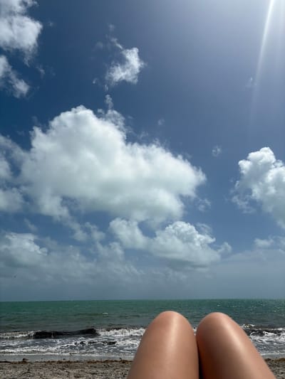 The image shows a scene at a beach in Key Biscayne, United States of America. The view is oriented towards the sky, which is predominantly blue with scattered white clouds and a visible sun glare. Part of the ocean and beach are visible, with waves crashing onto the shore. In the right edge of the frame, there are a pair of legs.