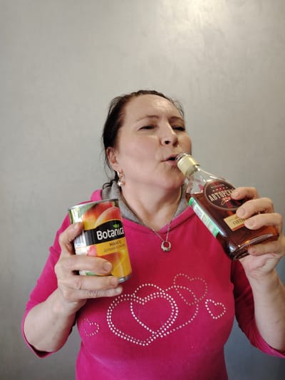 An adult woman is indoors against a plain gray wall. She is drinking from a bottle of what appears to be cognac and holding a can of "Botanica" brand mango fruit. The bottle is labeled "Авторский" (Avtorskiy). The woman is wearing a pink top with silver hearts. She has a silver necklace and earrings. The location is unspecified and cannot be verified as Neftekamsk, Russia, from the available image.