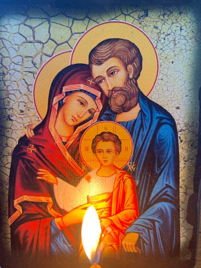 The image shows a religious icon depicting the Holy Family: Mary, Joseph, and the infant Jesus. Mary is on the left, with her arm around Joseph, who is on the right. The infant Jesus is in the center, held by Mary. All three figures have halos. The background is textured and appears aged, with cracks. In the foreground, a lit candle is obscuring part of the icon. The image is captured in Zelenodol'sk, Russia.