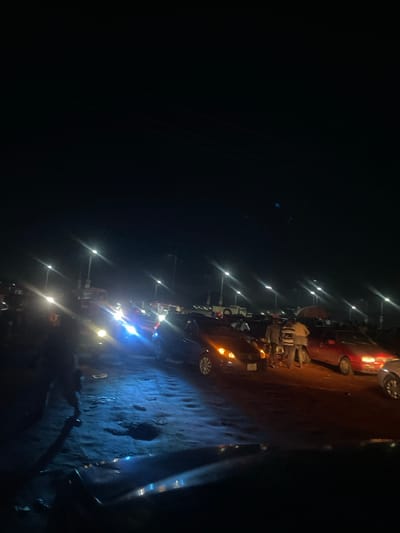 The image captures a nighttime scene on a dark, unpaved road in Sauka, Nigeria. The road surface is uneven and appears wet or muddy in places, reflecting the bright lights from passing vehicles and streetlights. A line of parked vehicles, including cars and what appear to be motorcycles or scooters, is visible along the side of the road. Some vehicles have their headlights on, illuminating the surrounding darkness.
The activity appears to be people gathered or interacting along the roadside. The presence of multiple vehicles suggests a potentially busy area or an event. The lighting is characterized by distinct, sharp streaks of light, indicative of a long exposure photograph or a camera struggling with low light conditions and bright light sources. The overall atmosphere is one of a typical nighttime street scene in a Nigerian locale, with the focus on the interplay of artificial lights and the dark environment. There are no discernible people clearly visible as distinct subjects, but their presence is implied by the parked vehicles and the general activity. The setting is clearly outdoors, on a road, under the cover of night.