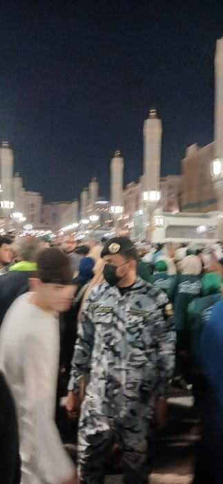 The image depicts a crowded nighttime scene outside Al-Masjid an-Nabawi (The Prophet's Mosque) in Medina, Saudi Arabia. In the mid-ground, a male subject is prominent, wearing a blue/grey digital camouflage uniform, a black beret adorned with a gold emblem, and a black protective face mask. His uniform features patches on the shoulders and chest with illegible text. He faces slightly to the right, looking forward. To his immediate left, partially obscuring the foreground, is a blurred individual wearing a light-colored long-sleeved garment and a dark head covering, also facing slightly right. Numerous other individuals, some wearing green tops and head coverings, are visible in the background, forming a dense crowd. The environment features multiple tall, light-colored minarets characteristic of Al-Masjid an-Nabawi, illuminated against a dark night sky. Various other mosque structures and streetlights providing ambient illumination are present in the blurred background. The ground appears to be a paved pedestrian area. The subjects are standing or walking, suggesting movement within the crowded outdoor space.