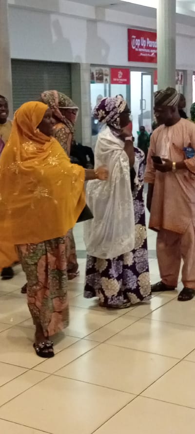 The image captures a scene indoors, likely a commercial establishment in Gandu Albasa, Nigeria. The lighting suggests an indoor setting, possibly a mall or a large shop, with tiled flooring. In the foreground, a woman wearing a vibrant yellow hijab and a patterned skirt is the central figure. She is engaged in conversation or gesturing towards another individual. To her right, another woman is dressed in a white hijab and a patterned dress with purple and white motifs. Further to the right, a man in a light pink traditional outfit is visible, holding a smartphone and looking downwards. The background shows a section of a shop with a red sign that reads "Top Up Parar" and what appears to be an entrance or a display. The overall atmosphere seems to be one of social interaction in a public space.