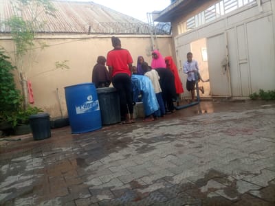 A group of people, including women in hijabs and a young man in a light blue shirt, are gathered outdoors in a paved area with wet cobblestones. A large blue water tank, a black bin, and a small tree are also visible. In the background, a building with a corrugated metal roof and a white metal gate is present. The scene appears to be in Wuro Dole, Nigeria. Some individuals are interacting with each other, with one person appearing to pour water from a blue pipe. The lighting suggests it is either early morning or late afternoon.