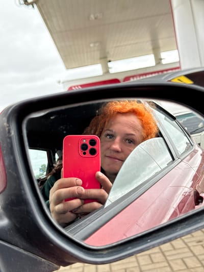 A person with bright orange hair is taking a selfie in the side mirror of a car. They are holding a red iPhone in their right hand, which is positioned in front of their face. The person appears to be in a car, as indicated by the reflection of the car's interior in the mirror. The background outside the car shows a gas station canopy and a cloudy sky. The person's face is looking directly at the camera, and they have a neutral expression. The image is taken from a low angle, looking up at the mirror. The car appears to be red. The ground is paved with interlocking bricks. The location is a gas station in Minderevo, Russia.