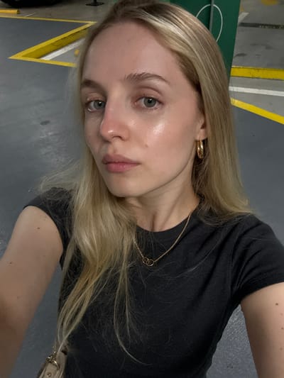 A close-up selfie of a blonde woman is taken in a dimly lit indoor parking garage. She is looking directly at the camera, with her head slightly tilted. The woman has fair skin, light blue eyes, and is wearing a black t-shirt, gold hoop earrings, and a delicate gold necklace. The background shows gray concrete flooring with yellow and white painted lines indicative of parking spaces. A section of a green sign with white markings is visible to the upper right of the frame. The overall lighting suggests it is nighttime or an underground location. The image does not provide any specific indicators of its location in Miami, United States of America.