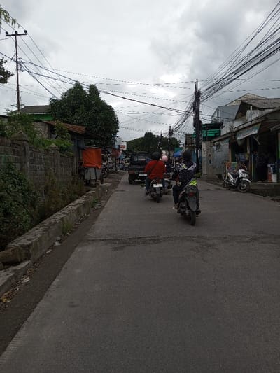 The image displays an overcast daytime street scene in the city of Melakudikadu, India. An asphalt road, exhibiting visible wear and cracks, occupies the foreground and recedes into the distance. On the left side, a concrete curb borders a shallow drainage ditch, behind which a low stone wall is partially covered by green vegetation. Further along the left, a stall-like structure with an orange tarp cover is present. On the right side, multiple low-rise buildings, appearing to be commercial establishments with storefronts, line the road. One building features a corrugated metal roof extension over its entrance, and a white scooter is parked on the sidewalk nearby. Overhead, numerous utility poles support a dense network of tangled electrical and communication wires that crisscross the sky. Vehicle traffic is observed moving away from the viewer. In the mid-ground, a black pickup truck is visible, followed by three motorcycles. The first motorcycle rider wears a red shirt and a dark helmet. The second motorcycle, positioned closer to the viewer, has a rider wearing a black and white patterned jacket with green accents and a dark helmet. A third rider with a light-colored helmet is partially visible between the two leading motorcycles.