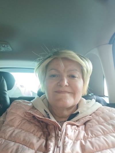 The image displays a head-and-shoulders portrait of an adult female, taken from a selfie perspective within a vehicle interior. The subject has short, light blonde hair and wears a light pink quilted jacket over a cream-colored hooded garment. Her gaze is directed straight at the camera, with a neutral facial expression. The vehicle's interior features a light-colored headliner, an overhead light console visible in the upper left, and a portion of a dark-colored seat headrest to the left. A bright light source, originating from an exterior window, is visible behind the subject. The location is Akulovo (Gp Kubinka), Russia.