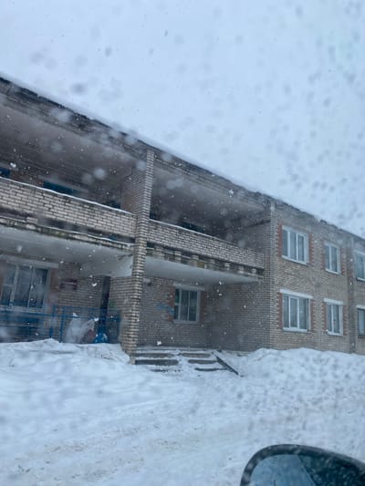 A snow-covered building is viewed from above. The facade is brick with rows of windows. A thick layer of snow covers the ground and roof, obscuring details. The perspective makes the building appear to rise vertically along the left edge of the image, with snow visible on both sides. Snow is falling, creating a blurred effect. Part of a vehicle mirror is visible on the right side.