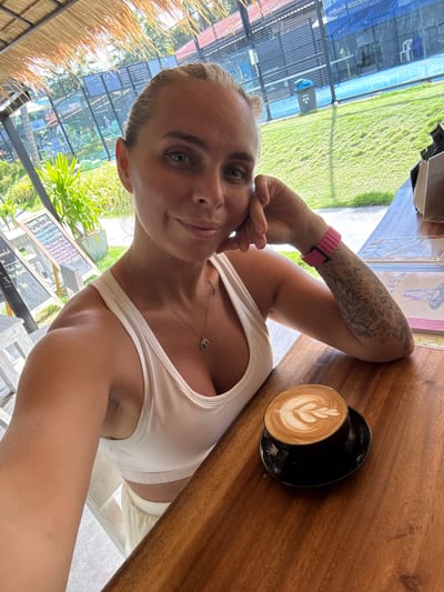 A woman with blonde hair tied back is taking a selfie while sitting at a wooden table. She is wearing a white tank top and has a tattoo on her left arm. A cup of coffee with latte art sits on the table in front of her. In the background, there are tennis courts visible, suggesting the location is a resort or sports complex in Baan Plai Laem, Thailand. A thatched roof is overhead, and lush greenery surrounds the area.