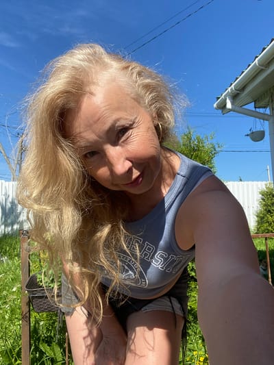 A blonde woman with fair skin and a light complexion, wearing a grey sleeveless crop top with "UNIVERSITY" written across it and denim shorts, is captured in a low-angle, selfie-style photograph outdoors under a clear blue sky. Her hair is styled in loose waves that fall across her face and shoulders. She is leaning forward, her face tilted towards the camera, with a slight smile. The background includes a white picket fence, green grass, bushes, and the corner of a white house with a gutter. The scene suggests a casual, sunny day in a residential area, likely in the city of Bol'shoe Zaborod'e, Russia.