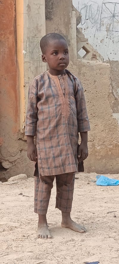 A young boy stands barefoot in a dusty environment, wearing a brown and blue plaid tunic and pants. His clothes are torn at the knees. He gazes off to the side with a somber expression. Behind him, there are crumbling, weathered buildings with peeling paint and holes in the walls, suggesting a dilapidated or impoverished area. A discarded blue plastic bag lies nearby.