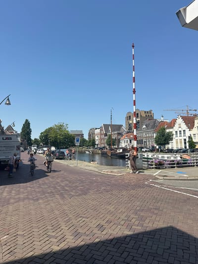 The image shows a canal in Zwolle, Netherlands, with a street running alongside it. The street is paved with brickwork, and there are several people on bicycles. A woman in a brown dress is standing near a red and white striped pole, which appears to be a traffic signal or barrier. Buildings line the opposite side of the canal, some of which have traditional Dutch architecture. A crane is visible in the background, suggesting ongoing construction. A white truck with "UTS HERSEVOORT VERHUIZINGEN" written on it is parked on the left side of the street. The sky is clear and blue, indicating a sunny day.