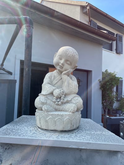 A concrete statue of a young meditating monk is positioned outdoors on a stone pedestal. The monk statue is seated cross-legged, with its right hand supporting its head as if in deep thought or sleep, and its left hand resting on a string of beads. The statue has a serene expression and is wearing traditional monk attire. The pedestal is light gray and textured with a pattern of irregular white spots, resembling a distressed or weathered finish.
Behind the statue, a building with light gray walls and brown shutters on the windows is visible. A dark-colored door is also present on the building. To the right of the statue and building, a green tree in a pot adds a touch of nature to the scene. The sky above is a clear blue. The overall environment suggests an outdoor setting, possibly a garden or courtyard, with a residential or commercial building in the background. The image exhibits lens flare with rainbow-like refractions, particularly noticeable in the upper left and lower portions. The location is identified as Gerstheim, France.