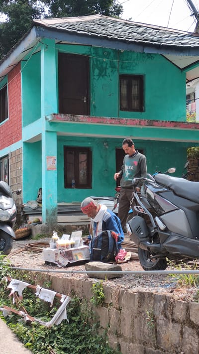 Two men stand outside a teal and brick building in Dharamkot, India. The building is two stories high with dark brown doors and windows. One man, wearing a grey shirt, looks down at another man who is dressed in a blue jacket, white hat, and red cap. The man in the blue jacket appears to be examining items placed on newspaper. A grey motorcycle is parked in front of the building. Debris, including newspaper and plastic bags, is scattered on the ground near vegetation. A stone stairway leads upward to the right of the scene.