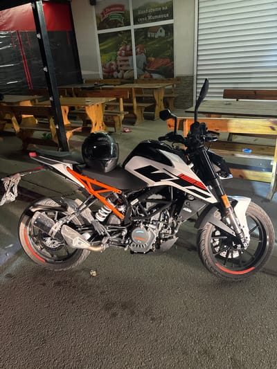 A white and black KTM 125 Duke motorcycle is parked on a dark asphalt surface, next to a building with wooden structures and a sign in Bulgarian. The sign reads "Колбасите на село Житница" in Cyrillic, translating to "Sausages of Zhitnitsa village." The location is likely in Mihailovo, Bulgaria. A black helmet hangs on the side of the motorcycle. The lighting appears to be artificial, possibly from streetlights or nearby buildings.