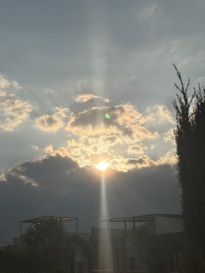 The image shows a view of the sky with bright sunlight shining through clouds. The sun is mostly obscured by the clouds, but beams of light radiate outwards. Part of a building is visible on the right edge of the frame, with some palm fronds partially obscuring it. Some tree branches are at the top of the frame. The photo appears to have been taken in San Juan Cuautlancingo, Mexico, as specified.