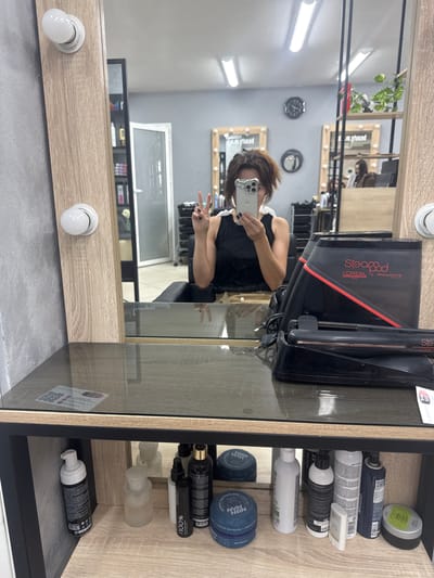 A person is taking a selfie in a salon mirror. They are wearing a black top with white trim and holding up a smartphone. The person is making a peace sign with their right hand. In the background, there's a reflection of the salon with chairs and other salon equipment. On a shelf to the right, there are various hair products, including bottles, cans, and jars. The room appears to be well-lit with fluorescent lights and vanity bulbs around a mirror. The setting is likely a professional hair salon.