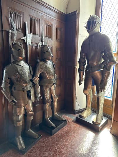 The image depicts three full suits of armor displayed at Reichsburg Cochem, a castle located in Cochem, Germany. Two suits are mounted on a dark brown wooden wall. Both are displayed lying down. Polearm weapons of medieval design are mounted along with the lower two suits of armor. The third suit is mounted horizontally above them, placed on a wall beneath a window. All three suits appear to be made of polished metal.