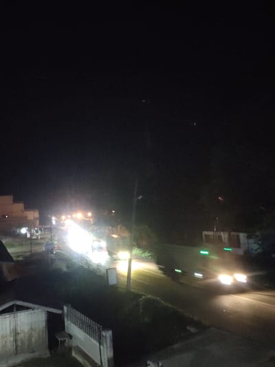 The image captures a nighttime scene on a road in Meunasahaluedrienlhoksukon Barat, Indonesia. A street or road curves through the frame, illuminated by the headlights of passing vehicles and streetlights. Several cars are visible, with their lights creating streaks due to motion blur, indicating they are in transit. Buildings are present on the left side of the road, with some showing lights within. The overall atmosphere is dark, typical of a rural or suburban area at night, with the artificial lights providing the primary illumination. The foreground shows what appears to be a low structure, possibly a shed or a small building, and a fence or railing. Vegetation is visible along the sides of the road and in the background, appearing as dark masses against the night sky. The presence of streetlights and moving vehicles suggests a functional road system within the locality.