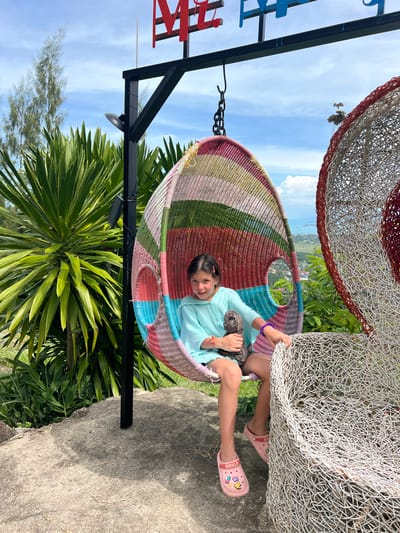 A young girl with dark hair is sitting in a colorful woven hanging chair at an outdoor viewpoint in Baan Thurian, Thailand. The chair is suspended from a black metal frame and features stripes of pink, red, green, and blue. The girl is wearing a light blue hooded top and pink clogs, one of which has cartoon charms. She is holding a dark gray stuffed animal. To the right of the hanging chair is a large, white, basket-like sculpture. Behind the girl, lush green tropical plants and trees are visible. In the distance, a town with buildings and a body of water can be seen under a partly cloudy sky. The ground in the foreground is a rough, gray concrete surface.