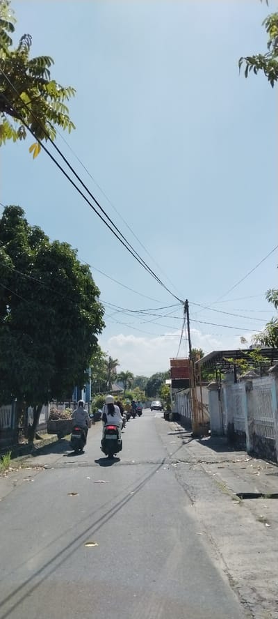 In Mataram, Indonesia, on a sunny day, people are riding scooters down a paved road. A large, leafy tree is on the left side of the road, and a white fence runs along the right side. Utility poles and wires are visible above the road and trees. Buildings line the street, some with signs. The sky is clear and blue.