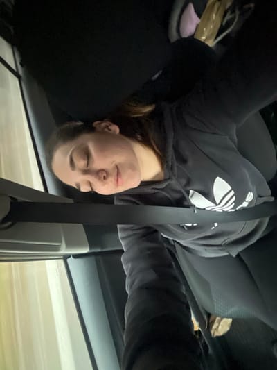 This image depicts a young adult female, oriented horizontally across the frame with her head towards the left. She appears to be asleep or unconscious, with her eyes closed and a neutral facial expression. She is wearing a grey hooded sweatshirt featuring a white Adidas trefoil logo on the chest and dark legwear. A black seatbelt is fastened across her torso and lap. She is seated in a dark-colored vehicle interior, likely a passenger seat. Through the window to her left, an out-of-focus background with horizontal motion blur indicates the vehicle is in motion. A light-colored, possibly crinkled object is partially visible on the floor or an adjacent seat to the right of the subject. The overall lighting within the vehicle is dim. The scene is located in Lonate Pozzolo, Italy.