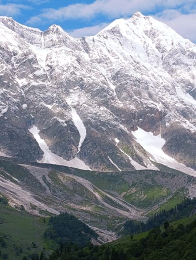 The image depicts a majestic snow-capped mountain range under a partly cloudy blue sky. The foreground transitions from a dense, dark green forest to a more sparsely vegetated, rocky slope with patches of snow. Further up the mountain, scree slopes and glaciers are visible, with white snow dominating the higher peaks and jagged rock formations breaking through. The overall scene is one of rugged natural beauty and a vast, untamed landscape. There are no people or man-made objects clearly discernible in the image.